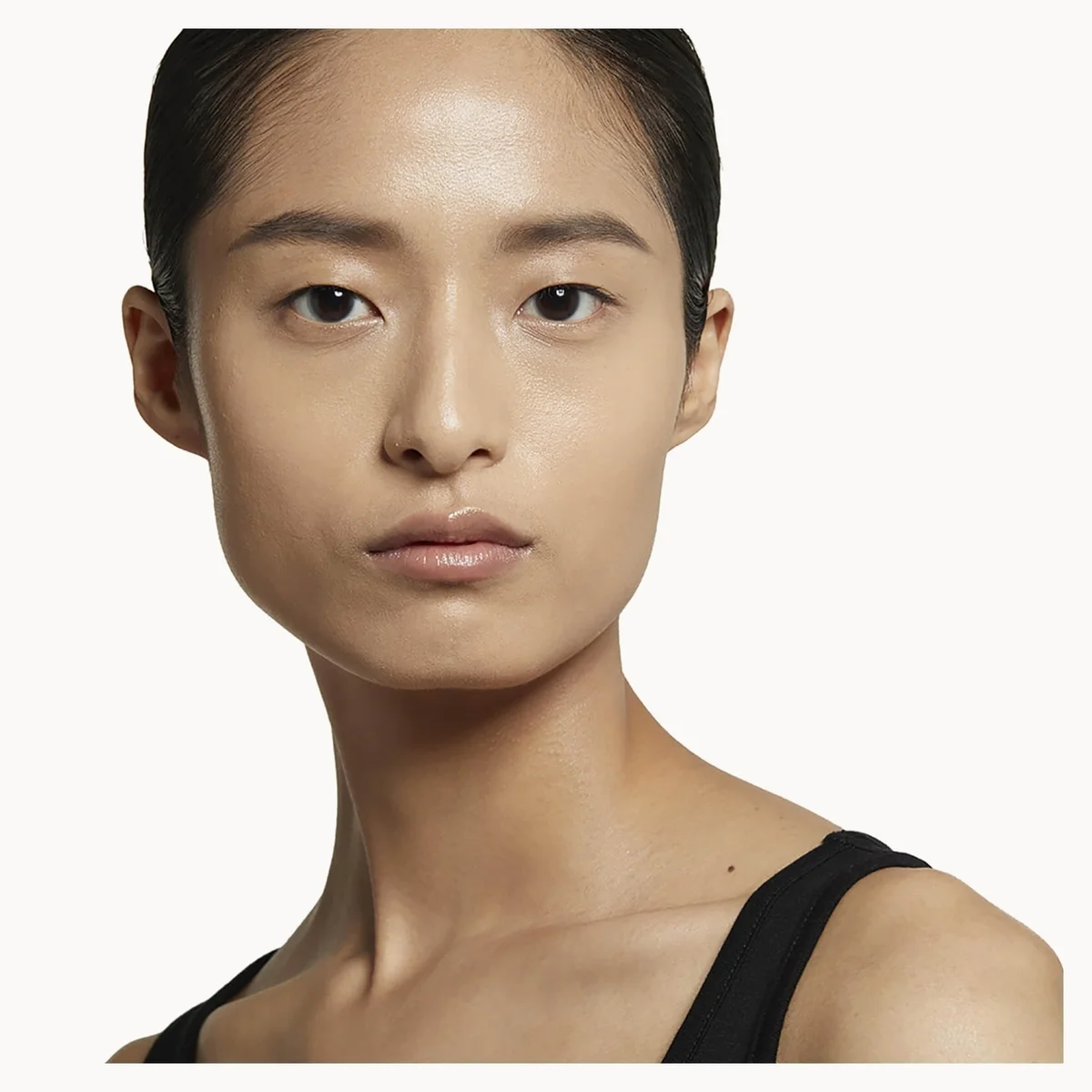 A close-up portrait of a model with dark hair pulled back, wearing a black spaghetti-strap top against a plain off-white background.