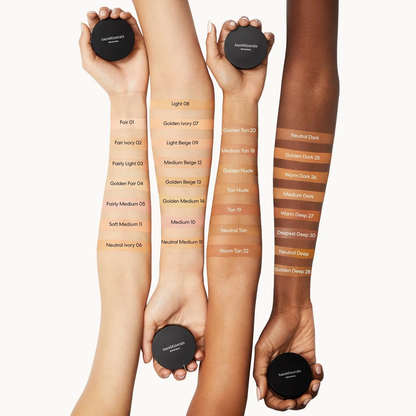 Four human arms and hands holding round black makeup compacts against a light gray background, with vertical swatches of foundation or tint labeled on the arms showing different shade options.
