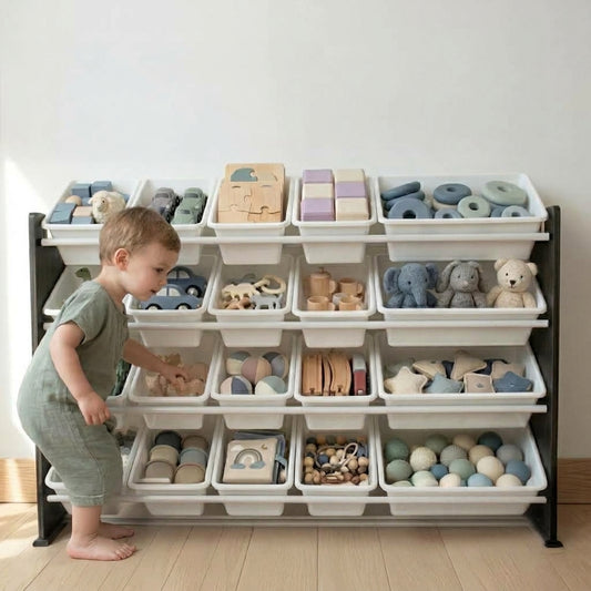 A toddler in a muted green outfit stands beside a large black-framed storage organizer filled with wooden blocks, balls, plush toys, and other baby toys in soft pastel colors, set against a plain light wall and wood floor.