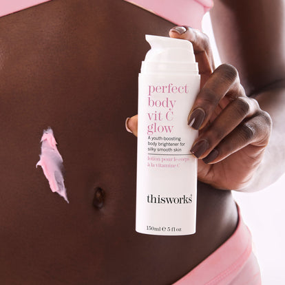 A person holding a white pump bottle labeled 'perfect body vit C glow' and 'thisworks' against their bare torso, with pink body cream visible on the skin.